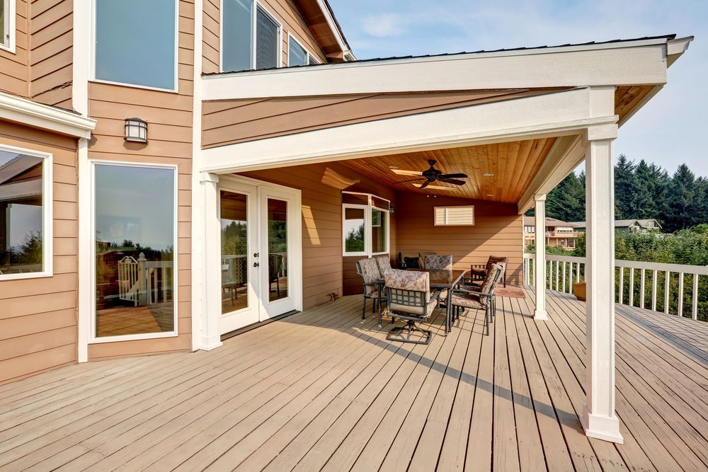 Wooden deck with outdoor furniture under a covered patio attached to a house with large windows.