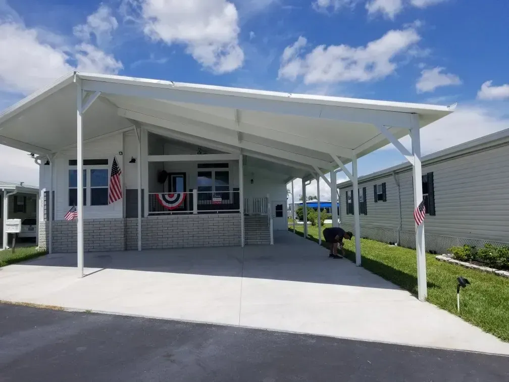 White carport with angled roof, covering the driveway of a light-colored mobile home, blue sky.