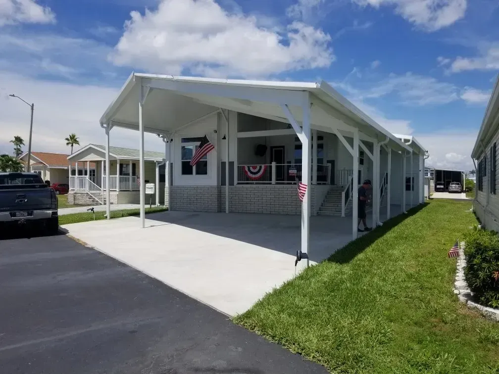 White manufactured home with carport, American flag, blue sky.
