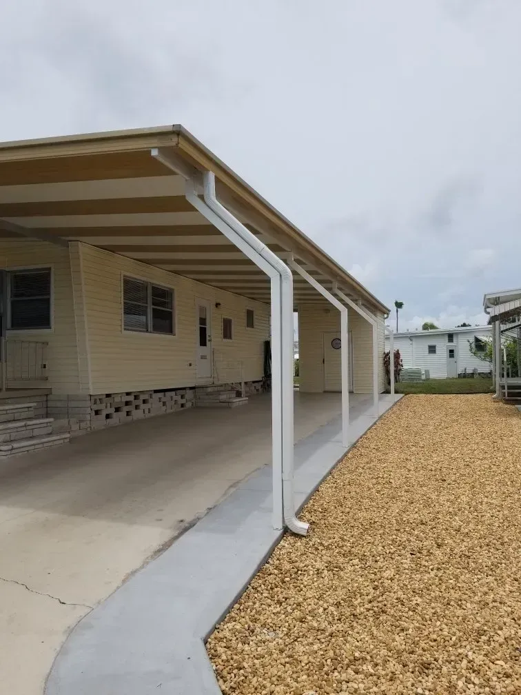 A beige mobile home with a carport. Concrete driveway and tan gravel border the side. Cloudy sky.