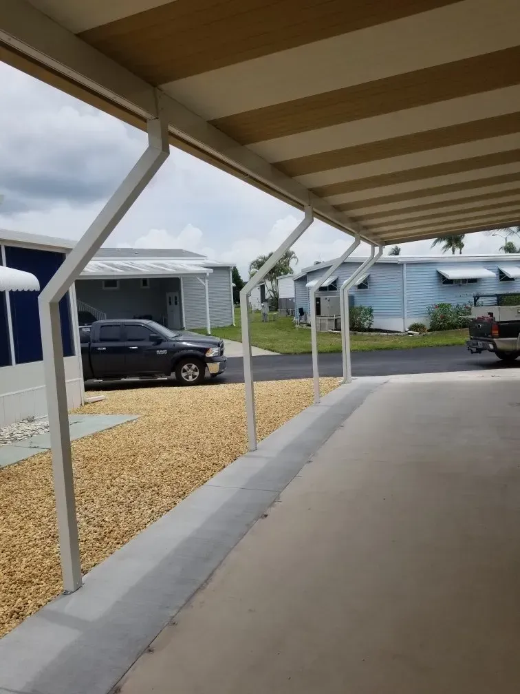White carport with tan and white striped roof over a concrete driveway lined with small tan rocks.