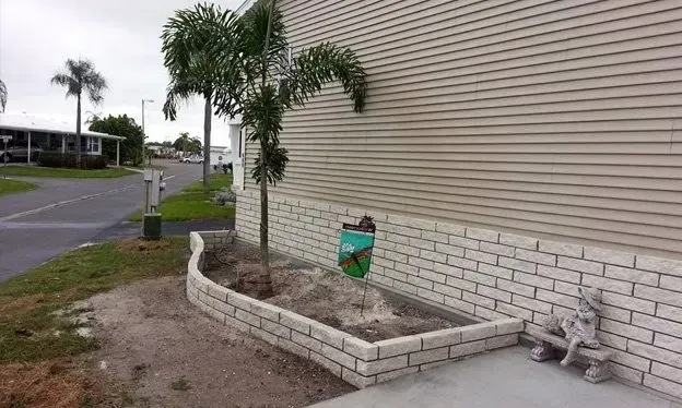 Palm tree in a brick-lined garden bed beside a light brown mobile home with a small statue and mailbox in a neighborhood.