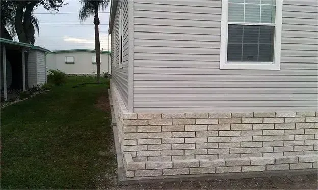 Corner of a light gray vinyl-sided house with a brick foundation, window, and grass.