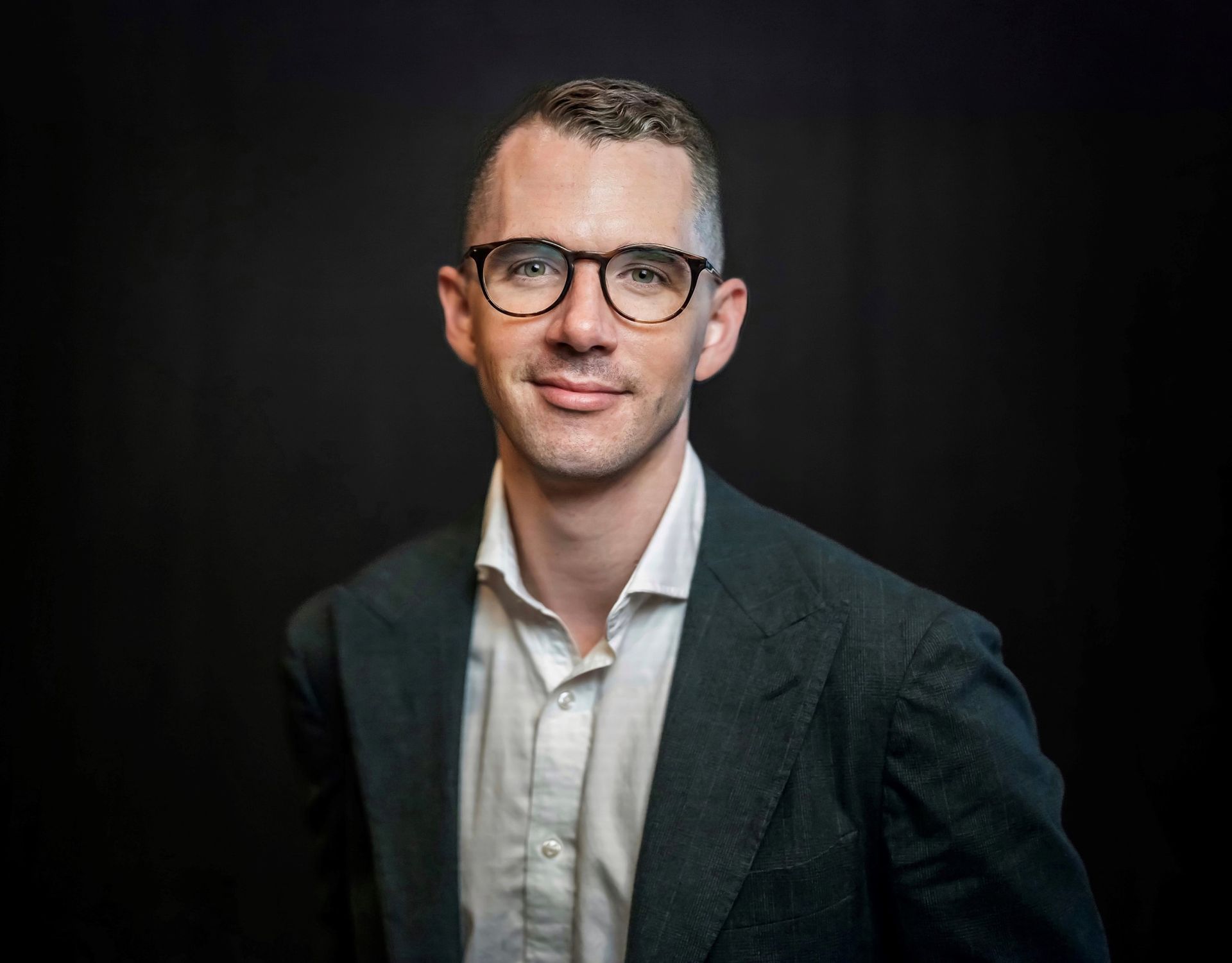 A man wearing glasses and a suit is standing in front of a black background.