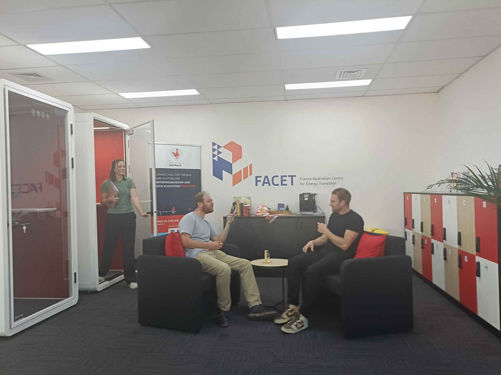 Two men seated on a sofa and talking, while a woman stands in a phone booth in an office.