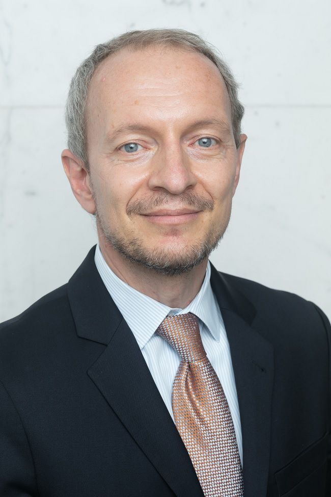Man in a suit and tie, smiling slightly, against a light-colored wall.