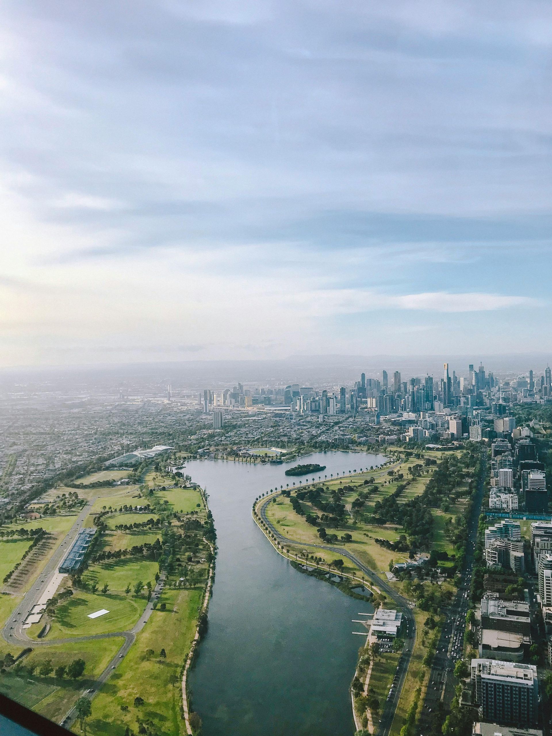 Aerial view of a city with a lake in the foreground, green fields and buildings, under a cloudy sky.