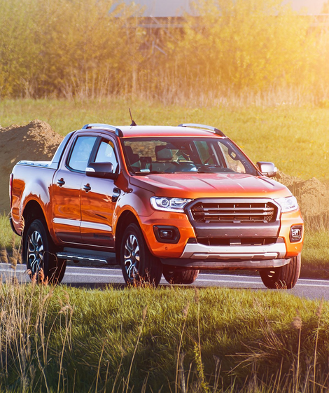 A bright orange pickup truck driving on a paved road through a grassy field during a sunny day.