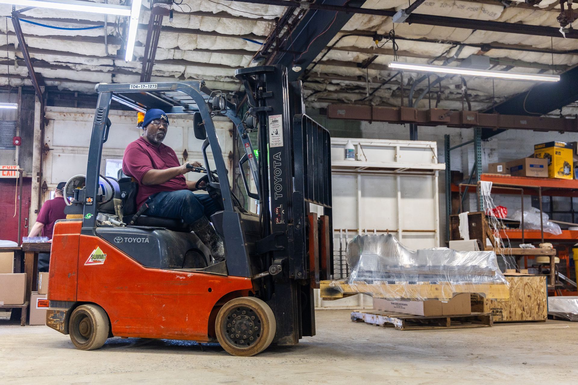 A person wearing a beanie drives an orange forklift inside a warehouse, carrying a pallet wrapped in plastic.