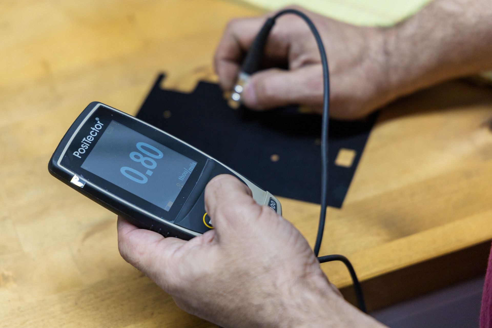 A person using a handheld PosiTector gauge to measure the coating thickness on a black metal plate.