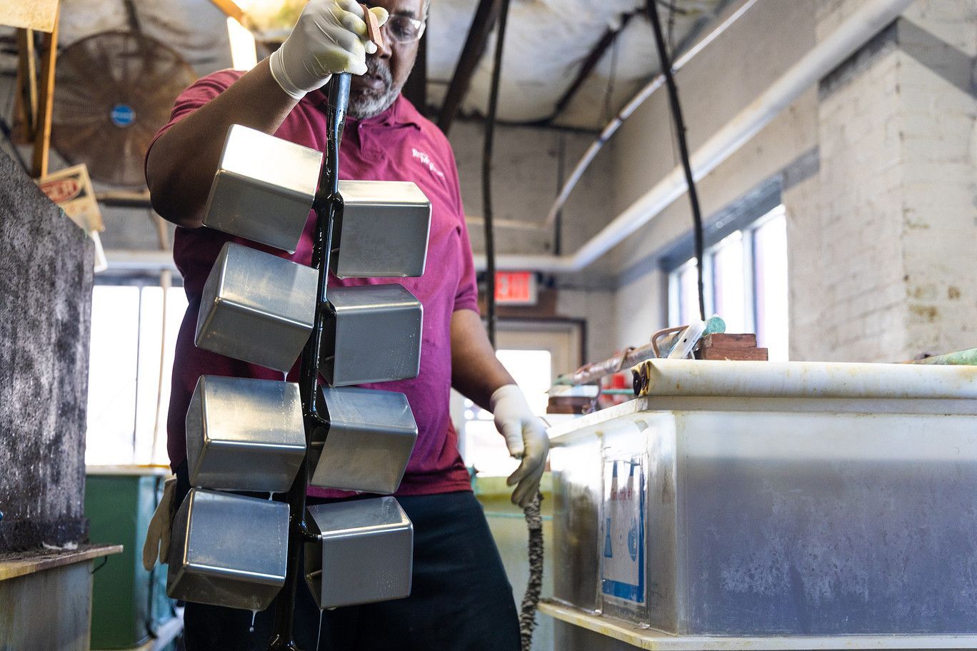 A person in a maroon shirt and safety gloves holds a vertical rack of eight metal cubes over an industrial fluid tank.