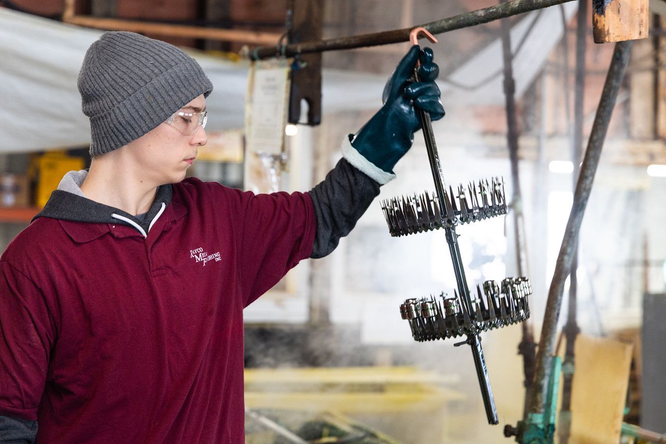A person wearing protective eyewear and gloves holds a metal rack of small parts near steam in an industrial workshop.
