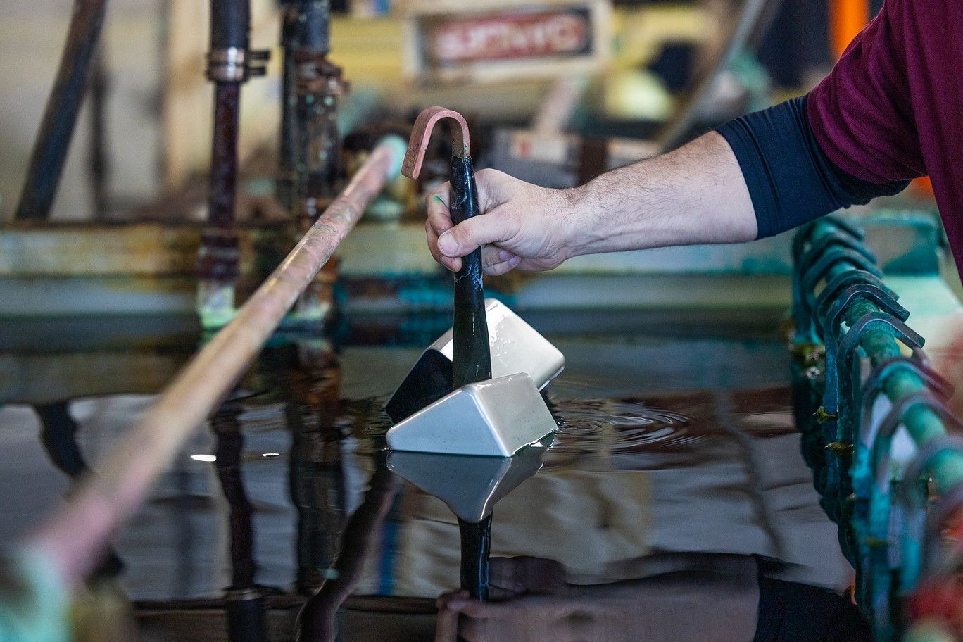 A worker dips a metallic, triangular part into a dark liquid bath in an industrial setting.