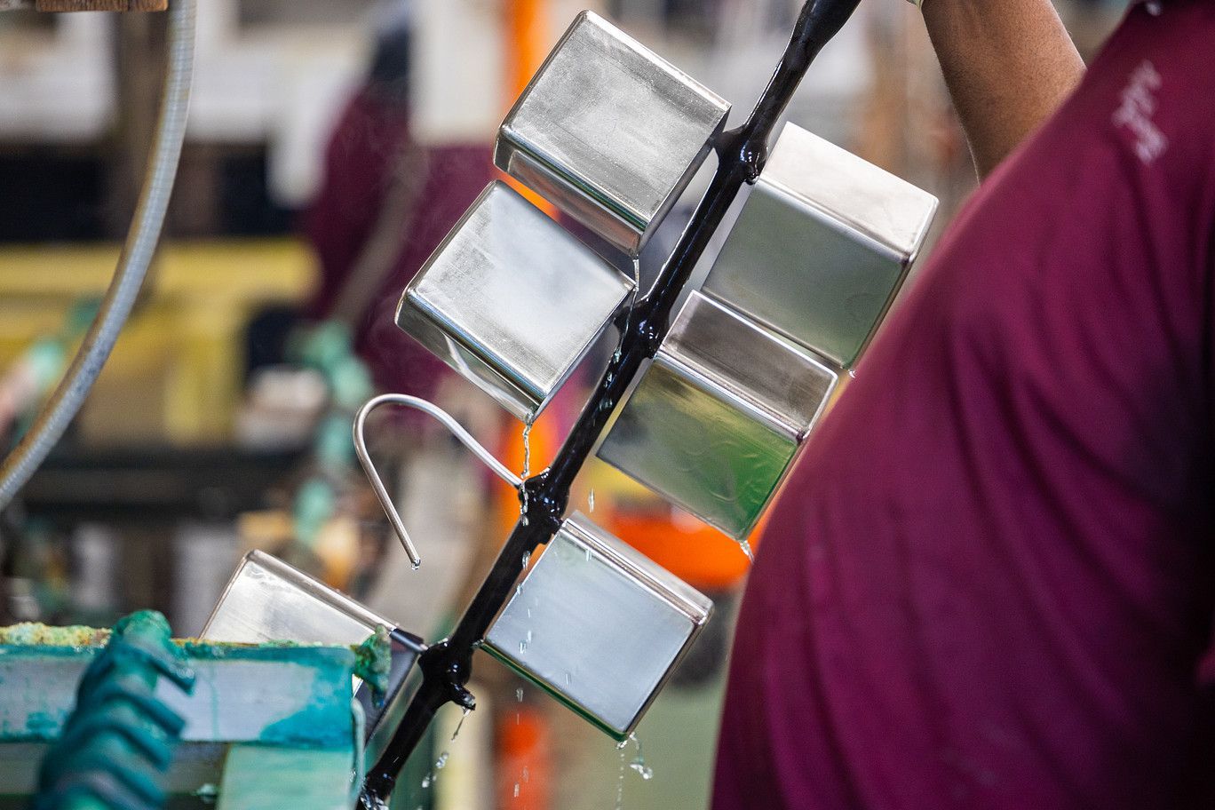 A person in a maroon shirt holds a metal rack of shiny, rectangular containers dripping water in an industrial setting.