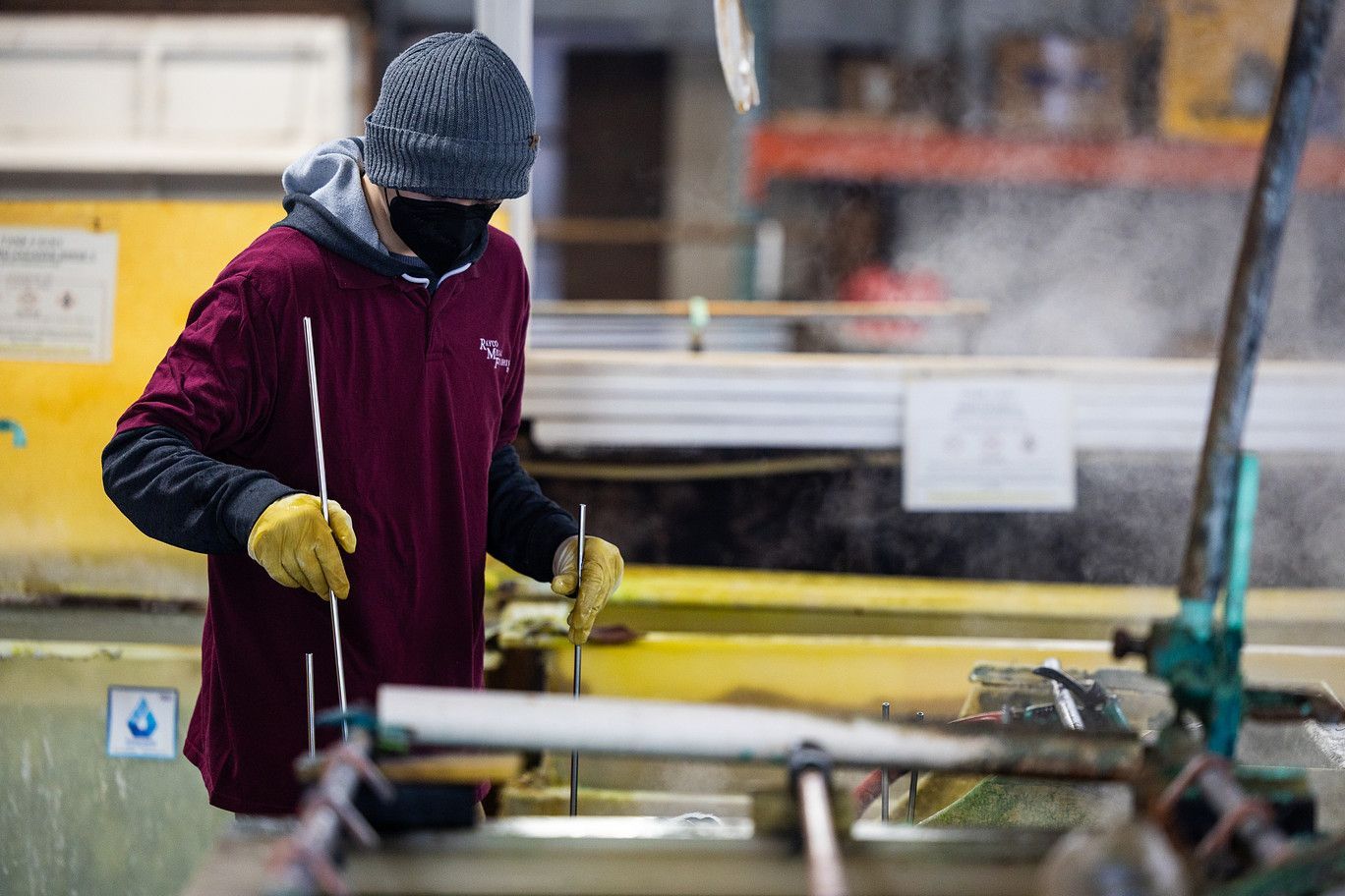 A person wearing a beanie, mask, and yellow gloves holds thin rods over a steaming industrial vat in a workshop.