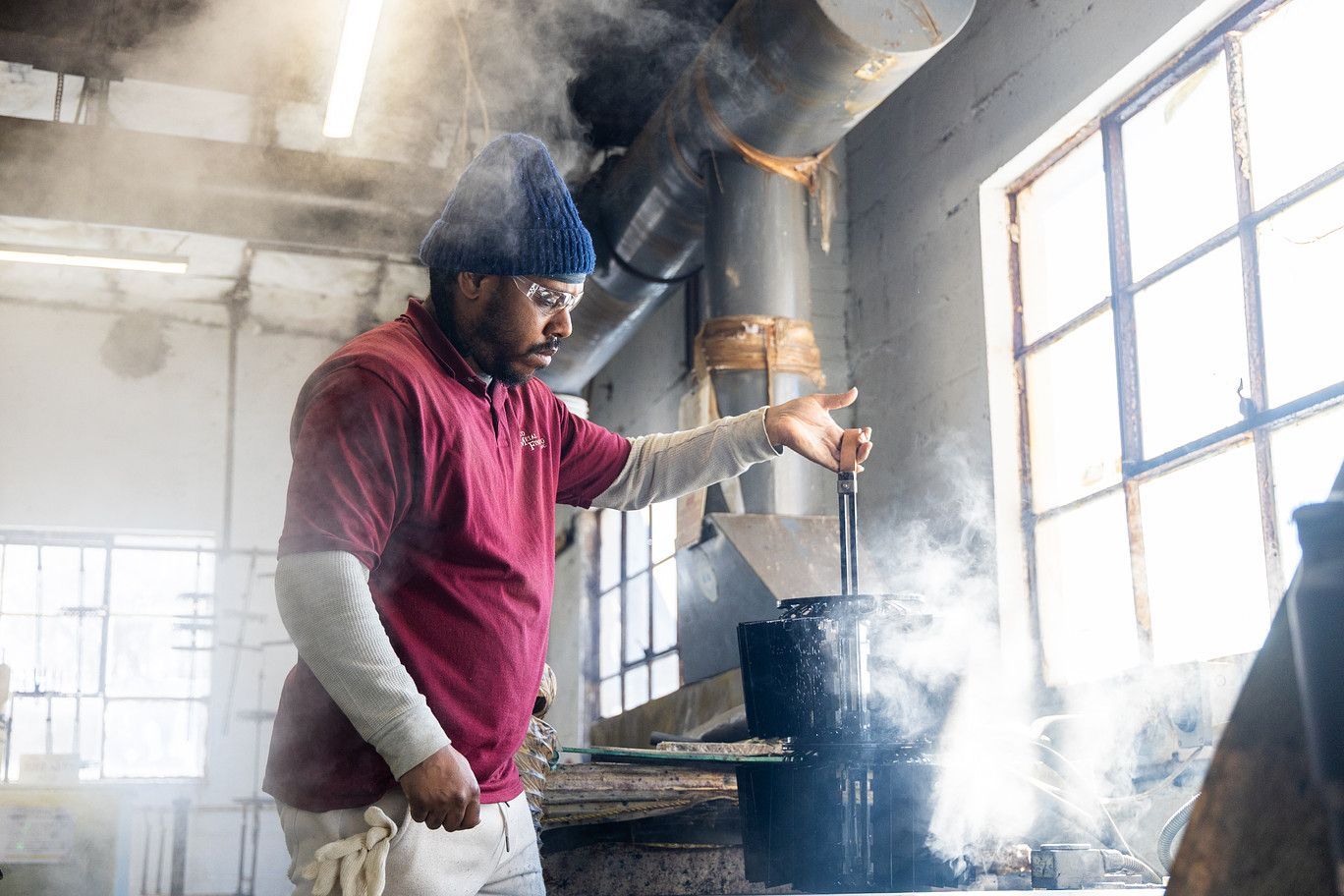 A person in a red polo and beanie works in a smoky industrial workshop, stirring a dark vessel with a metal tool.