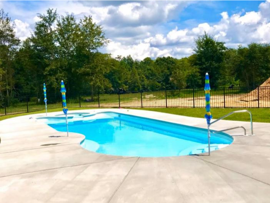 A blue backyard swimming pool with a concrete deck, two umbrellas, and a black fence, surrounded by green trees.
