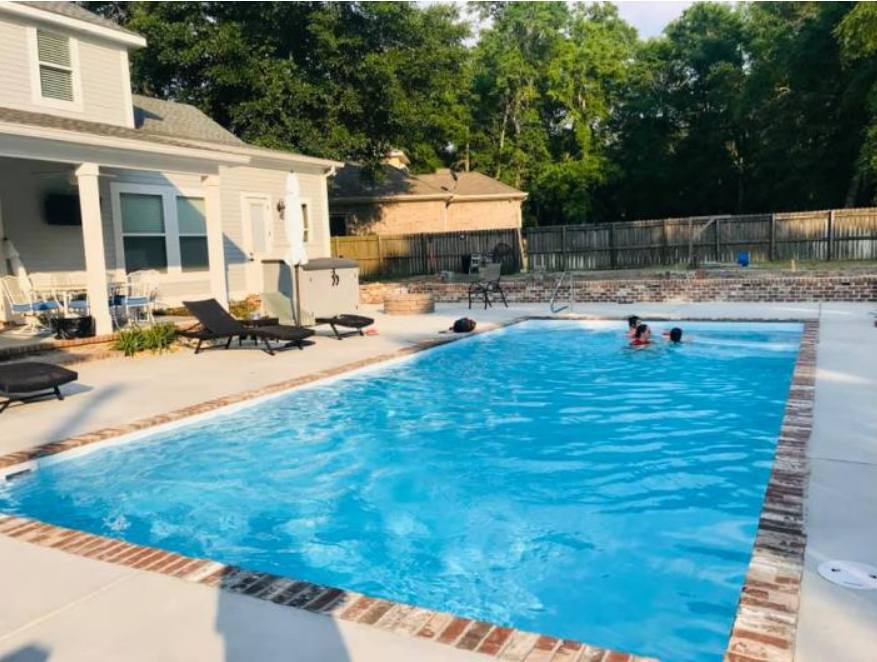 People swimming in a bright blue rectangular pool at a home with a patio, lounge chairs, and a wooden fence.
