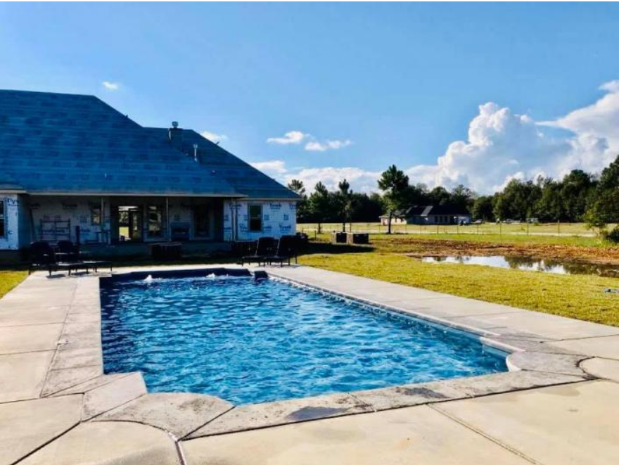 A rectangular swimming pool with blue water sits on a concrete patio in front of a house on a sunny, grassy day.