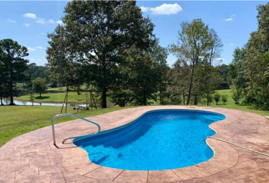A kidney-shaped swimming pool with blue water and tan stone decking overlooks a grassy yard, trees, and a pond.
