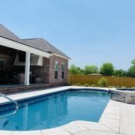 A blue swimming pool with an attached spa, situated next to a brick house with a covered patio under a clear blue sky.