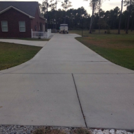 A long concrete driveway leading toward a brick house with a boat parked in the background.