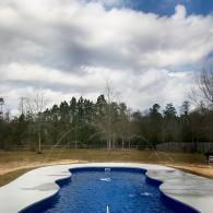 An outdoor, blue-tiled swimming pool surrounded by a concrete deck with a forest and a cloudy sky in the background.