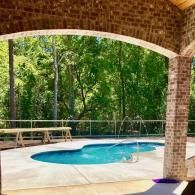 A kidney-shaped swimming pool sits on a patio under a brick-arched pavilion, surrounded by lush green trees.