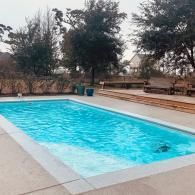 A rectangular swimming pool filled with bright blue water, surrounded by concrete decking, trees, and wooden patio furniture.
