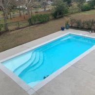 A rectangular backyard swimming pool with a shallow-end staircase, surrounded by light gray concrete, outdoors.