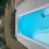 A rectangular, light blue swimming pool set in a backyard patio area next to a grassy lawn.