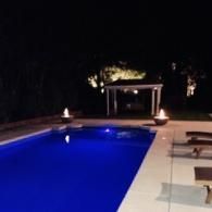 A swimming pool at night with two fire bowls on the deck and a small, illuminated gazebo in the background.