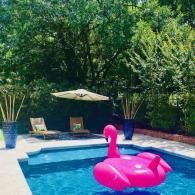 A bright pink flamingo pool float in a blue swimming pool with lounge chairs and an umbrella on the patio behind it.