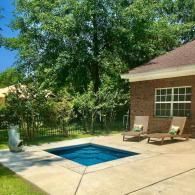A small, rectangular, blue-tiled swimming pool sits on a concrete patio beside a brick house with two lounge chairs.
