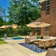 A tan sectional sofa and umbrella sit on a blue patterned rug on a concrete patio beside a small rectangular pool.