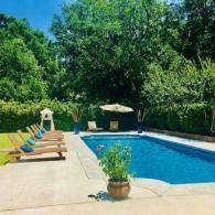 A blue swimming pool surrounded by a stone deck, wooden lounge chairs, a patio umbrella, and lush green trees.