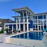 A modern two-story home with blue accents, white pillars, and a rectangular infinity pool on a sunny day.