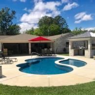 A backyard patio featuring a freeform swimming pool with a red umbrella, outdoor dining, and a stone covered pavilion.