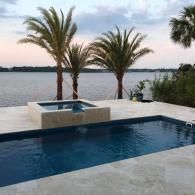 A rectangular swimming pool and raised hot tub on a stone patio overlooking a river at sunset with palm trees.