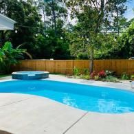 A kidney-shaped swimming pool with an attached spa, surrounded by a concrete deck and a wooden fence, under lush trees.