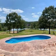 A bean-shaped, blue swimming pool set in a backyard with a stone patio, overlooking a lake and grassy area with trees.