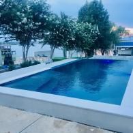 A rectangular swimming pool with deep blue water, surrounded by a light-colored concrete deck and flowering trees.