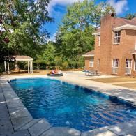 A rectangular swimming pool with blue water sits on a stone patio next to a red brick house and a covered pergola.