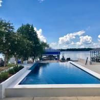 A rectangular swimming pool sits next to a calm lake under a bright blue sky with scattered clouds.