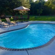 A backyard swimming pool with blue water and a brick-edged border, accompanied by patio furniture and an umbrella.