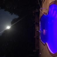 A bright moon illuminates the dark night sky above a glowing blue swimming pool at night.