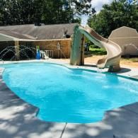 A light blue backyard swimming pool with a tan slide, a stone-walled house, and trees in the background.
