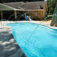 A kidney-shaped swimming pool with active water fountains on a sunny day in a backyard with a house in the background.