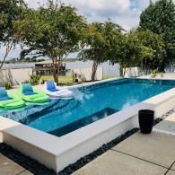 A rectangular swimming pool with blue water and inflatable loungers, positioned near a lake with trees and a dock.