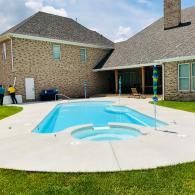 A rectangular swimming pool with an integrated spa, situated on a concrete patio in the backyard of a brick house.